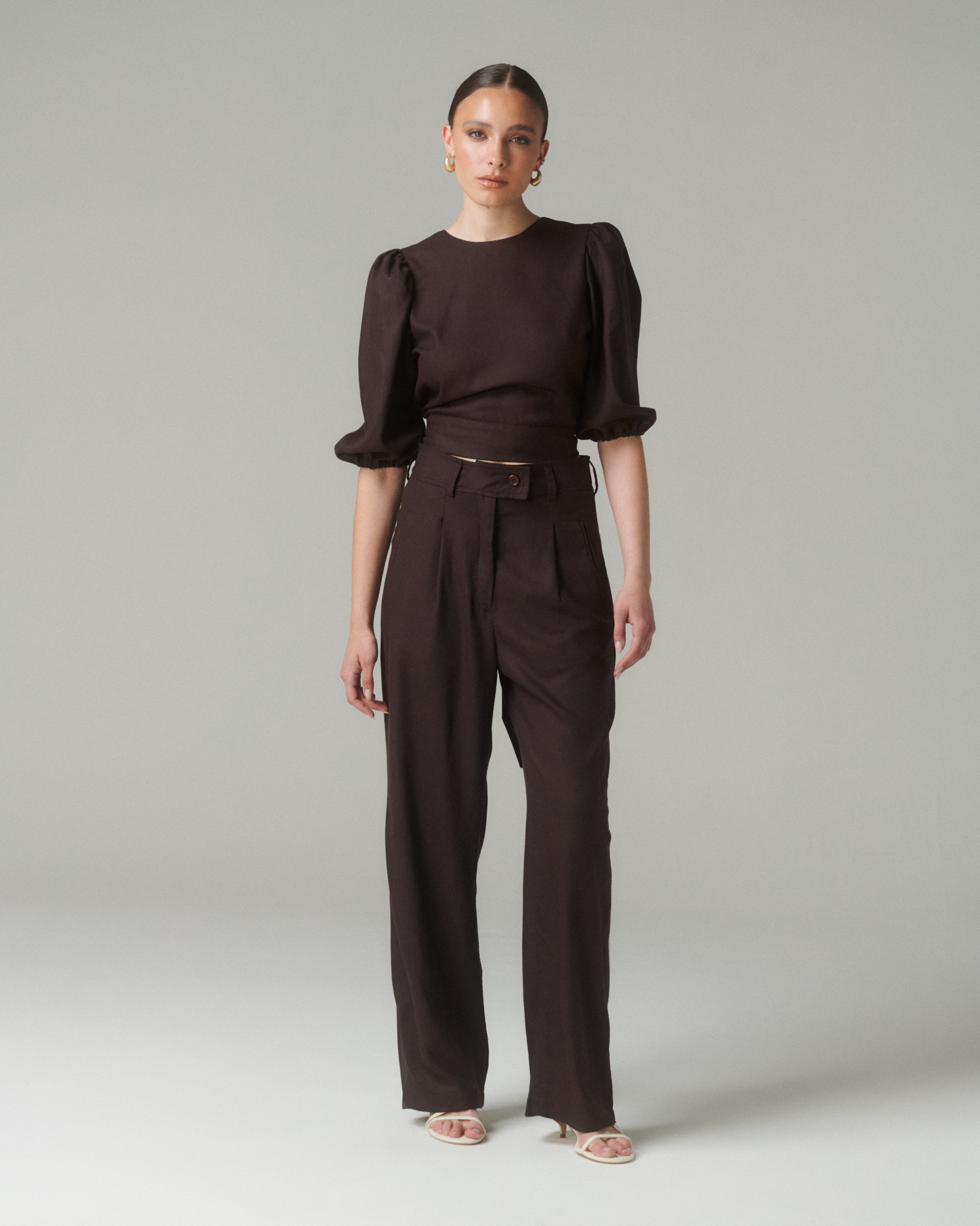 Vista frontal completa de la Blusa Aura Café con pantalones negros, destacando el cuello medio y las mangas abullonadas ¾.