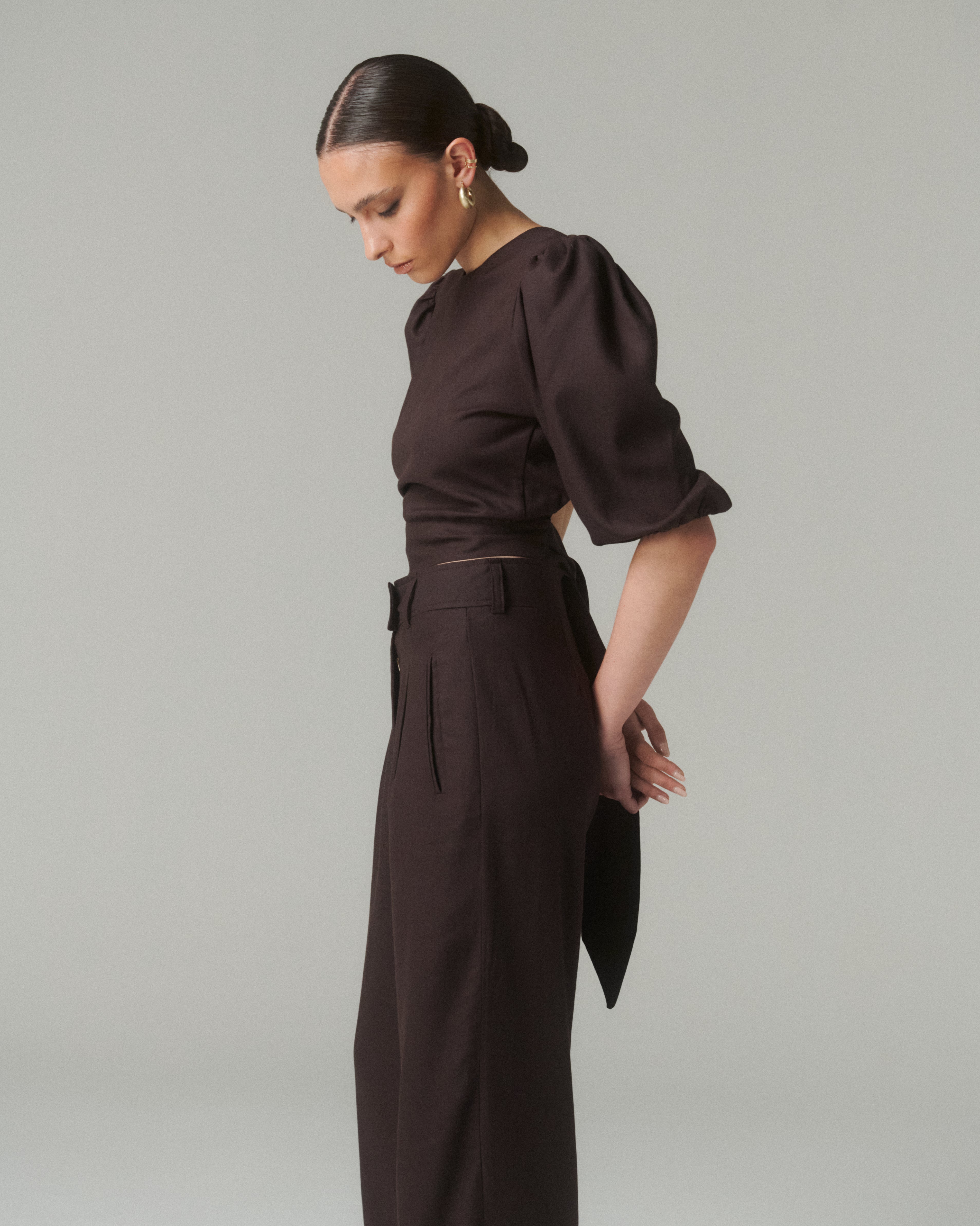 Vista lateral de la Blusa Aura Café, resaltando la caída de la prenda y el ajuste en la cintura con el lazo, combinada con pantalones negros.