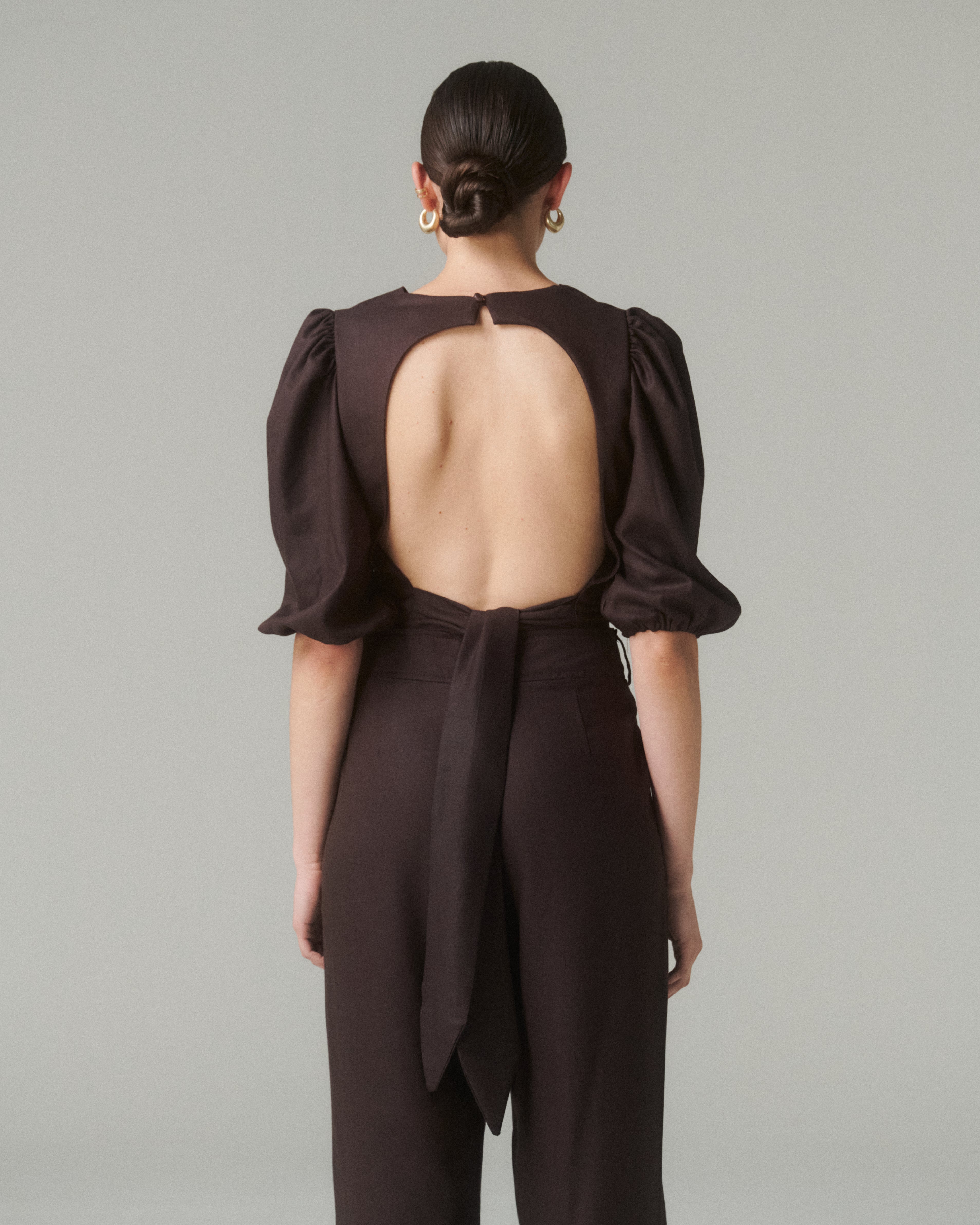 Vista trasera de la Blusa Aura Café, mostrando el corte en la espalda y el lazo ajustable, combinada con pantalones negros.