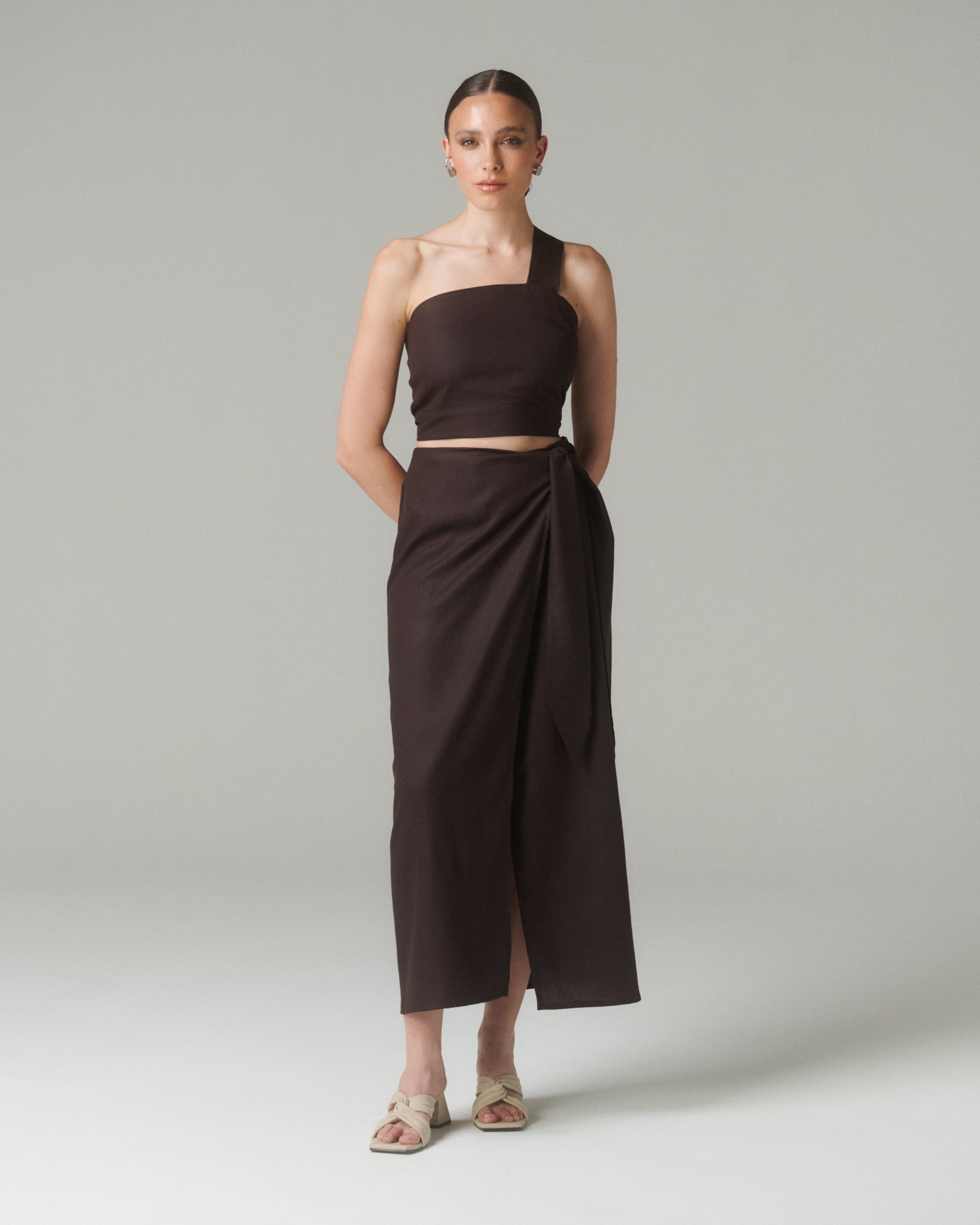 Modelo de pie con la Falda Lara Café y un top del mismo color o tono arena/tierra.  Fondo claro o beige para resaltar el tono café.  Poses naturales que muestren la caída fluida y el lazo lateral.
